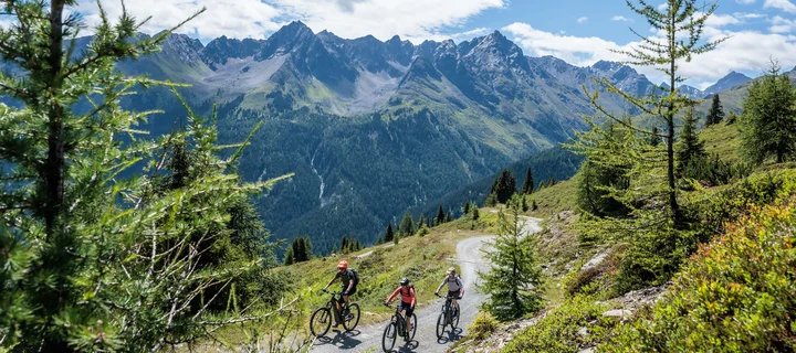 Ferienglück and Alpenland | Your stylish hotels in Ischgl Three mountain bikers ride along a trail with an alpine mountain backdrop