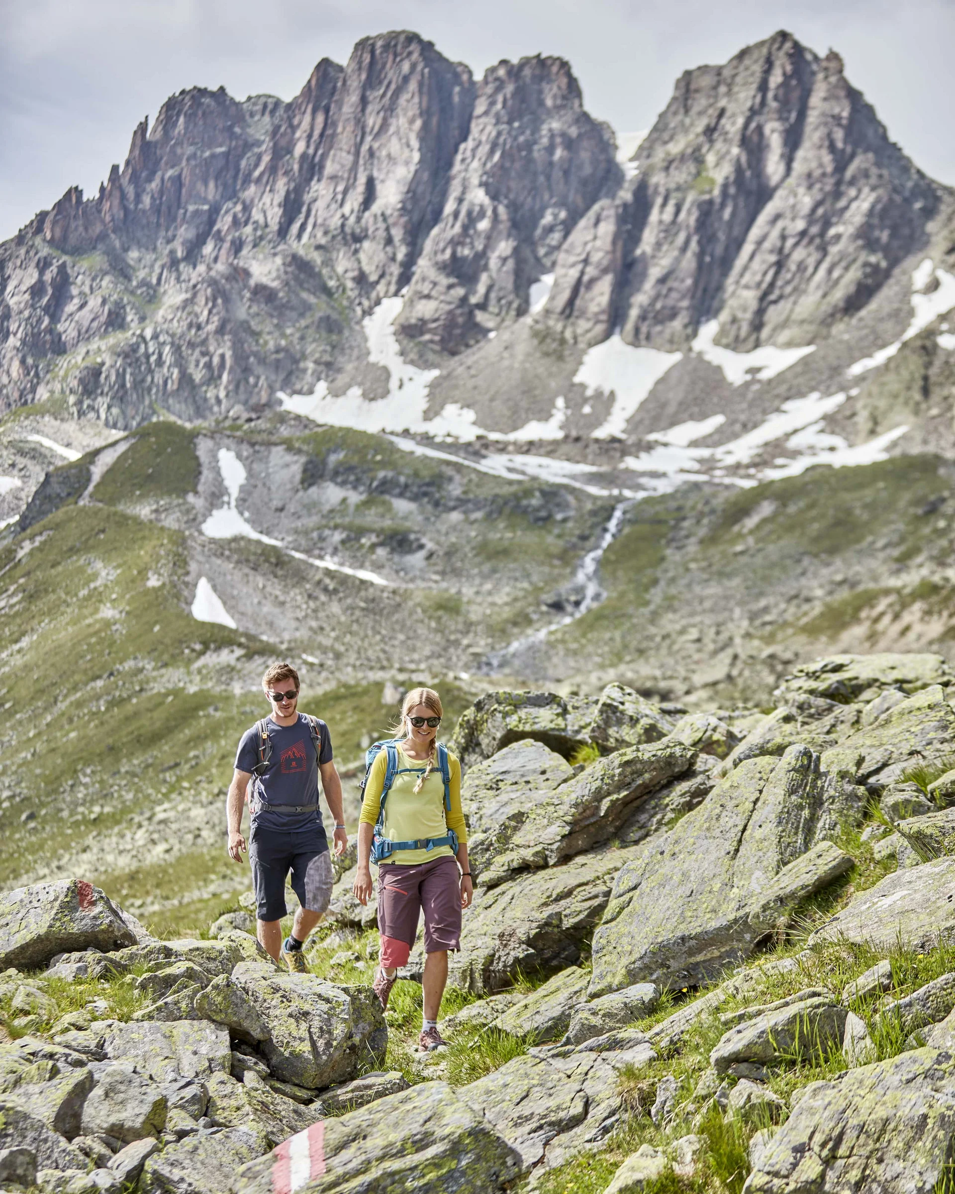 Ferienglück and Alpenland | Your stylish hotels in Ischgl Two hikers on a rocky mountain trail below rugged peaks with patches of snow